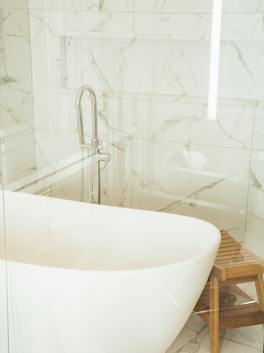 A white freestanding bathtub in a bright marble bathroom with a wooden stool