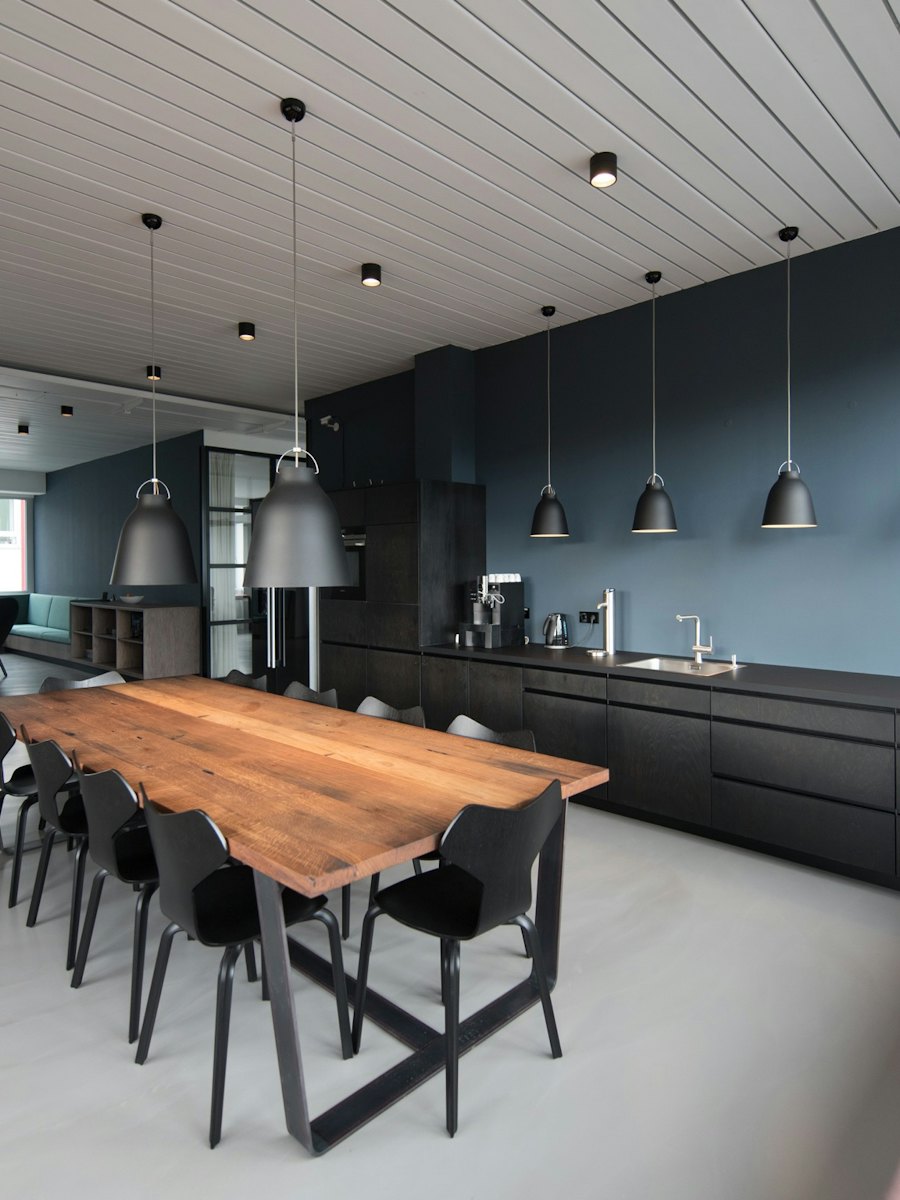 An office break room with a long wooden table, pendant lights and a clean kitchen
